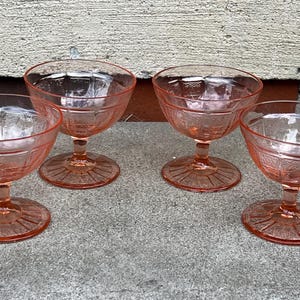 May include: Four vintage pink glass dessert or champagne coupe glasses. Each glass has a round base, a stem, and a bowl with a decorative pattern. The glasses are arranged on a gray surface.