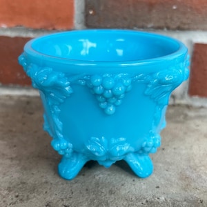 May include: A vibrant turquoise glass bowl with a detailed grape and leaf design. The bowl has three small feet and a rounded rim. The embossed pattern adds texture to the bowl's exterior, creating a vintage aesthetic.