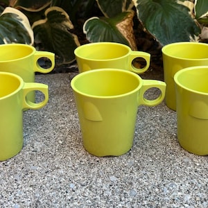 Könnte beinhalten: Sechs limettengrüne Tassen mit abgerundeten Griffen sind auf einer strukturierten Oberfläche angeordnet. Die Tassen haben ein leicht abgewinkeltes Design und ein kleines, erhabenes Detail an der Seite. Die Tassen bestehen aus einem festen, undurchsichtigen Material.