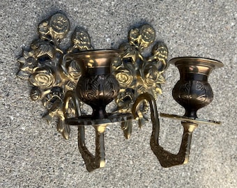 May include: A pair of ornate brass candle holders with floral designs. Each holder has a single candle cup and a decorative scrollwork arm.