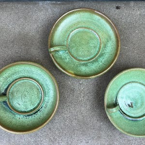 Op de afbeelding: Drie handgemaakte keramische theekopjes en schotels. De theekopjes en schotels hebben een gevlekte groene kleur met een gouden rand. De theekopjes hebben een klein handvat. De schotels hebben een verzonken midden om het kopje vast te houden.