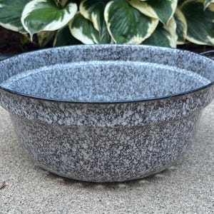 Może przedstawiać: Nakrapiana szara emaliowana miska z czarnymi uchwytami. Misa ma szeroki, płytki kształt i czarną krawędź. Nakrapiany wzór to mieszanka szarego i czarnego. Misa stoi na betonowej powierzchni.