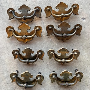 May include: Eight antique brass drawer pulls with a decorative, ornate design. Each pull features a curved handle and a floral-shaped backplate. The brass has a warm, aged patina, suggesting vintage or antique origins. These pulls are suitable for furniture restoration.