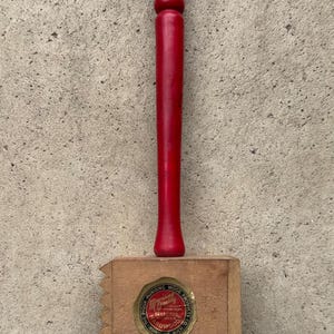 May include: A wooden meat mallet with a bright red handle and a natural light brown head. The head features a circular sticker with the text Munising Wood Products Co. The Piqua Munising Quality Selected Hardwood Munising Mich.