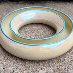 Peut inclure: Un récipient circulaire en céramique avec une base jaune pâle et une finition irisée et nacrée. L'objet présente un large anneau extérieur plat et un anneau intérieur plus petit et surélevé, tous deux bordés d'un fin liseré bleu sarcelle.