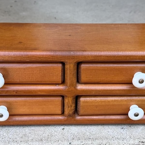 May include: A rectangular wooden storage unit featuring four drawers arranged in two rows. Each drawer is constructed from medium-toned wood and is equipped with a small, round, white ceramic knob centered on the front panel.