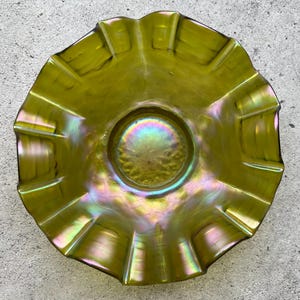 Puede incluir: Un cuenco de vidrio verde iridiscente con borde ondulado. El cuenco tiene una base circular y un borde acampanado y festoneado. El vidrio tiene un aspecto brillante, con toques de púrpura, rosa y dorado.