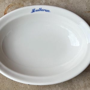 May include: White oval serving dish with a slightly raised rim. The word "Lutheran" is printed in blue script across the top. The dish is empty and appears to be made of ceramic.