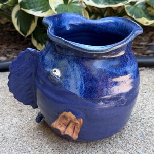 May include: A ceramic pitcher shaped like a fish with a deep blue glazed finish. The vessel features a wide, wavy rim, a prominent fin on the side, a single white eye, and a small, textured tan fin near the base. The surface shows variations in blue.