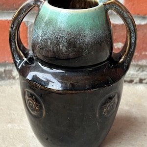 May include: A small, ceramic vase with two handles. The vase has a dark brown base with embossed circular designs. The top portion is a lighter brown with a turquoise interior. The handles are arched and attached to the top portion.