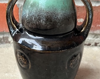 Può includere: Un piccolo vaso in ceramica con due manici. Il vaso ha una base marrone scuro con disegni circolari in rilievo. La parte superiore è marrone chiaro con un interno turchese. I manici sono arcuati e attaccati alla parte superiore.
