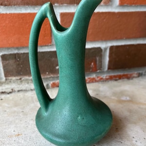 May include: A small, green ceramic pitcher with a curved handle and a flared spout. The pitcher has a rounded base and a textured surface. The background is a brick wall. This decorative pitcher is suitable for displaying flowers or as a decorative accent.