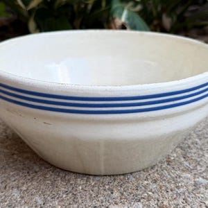 May include: A cream-colored ceramic mixing bowl featuring three distinct horizontal blue stripes encircling the upper exterior rim. The bowl has a wide, open top and a tapered base, showcasing a smooth, glossy finish with subtle signs of wear.
