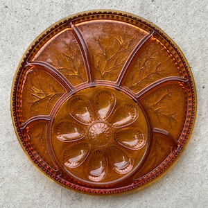 Peut inclure: Plat à servir vintage en verre ambré, avec un design divisé. Le plat rond présente une section centrale en forme de fleur entourée de six compartiments. Des détails décoratifs de feuilles et de bordure perlée rehaussent l'esthétique vintage. Une pièce élégante pour servir des collations.