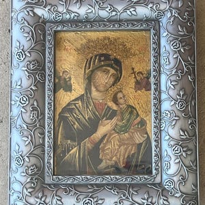 May include: A framed religious icon featuring a depiction of the Virgin Mary holding the Christ Child. The artwork has a gold background and is enclosed in a silver-colored frame with a floral design.