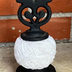 Puede incluir: Un portahilos de metal negro con una parte superior decorativa, que sostiene una gran bola de hilo blanco. El portahilos está sobre una superficie de piedra frente a una pared de ladrillo.