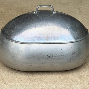 May include: An oval-shaped silver-colored metal roasting pan with a matching domed lid. The pan features two side handles and a single arched handle on top of the lid. The surface of the metal shows signs of wear with minor scratches and scuffs.