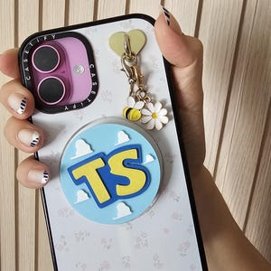 May include: A white smartphone case with a subtle floral pattern, a purple camera frame labeled CASETiFY, and a circular light blue grip featuring white clouds and the yellow letters TS. A gold heart charm hangs with a bee and a white flower.