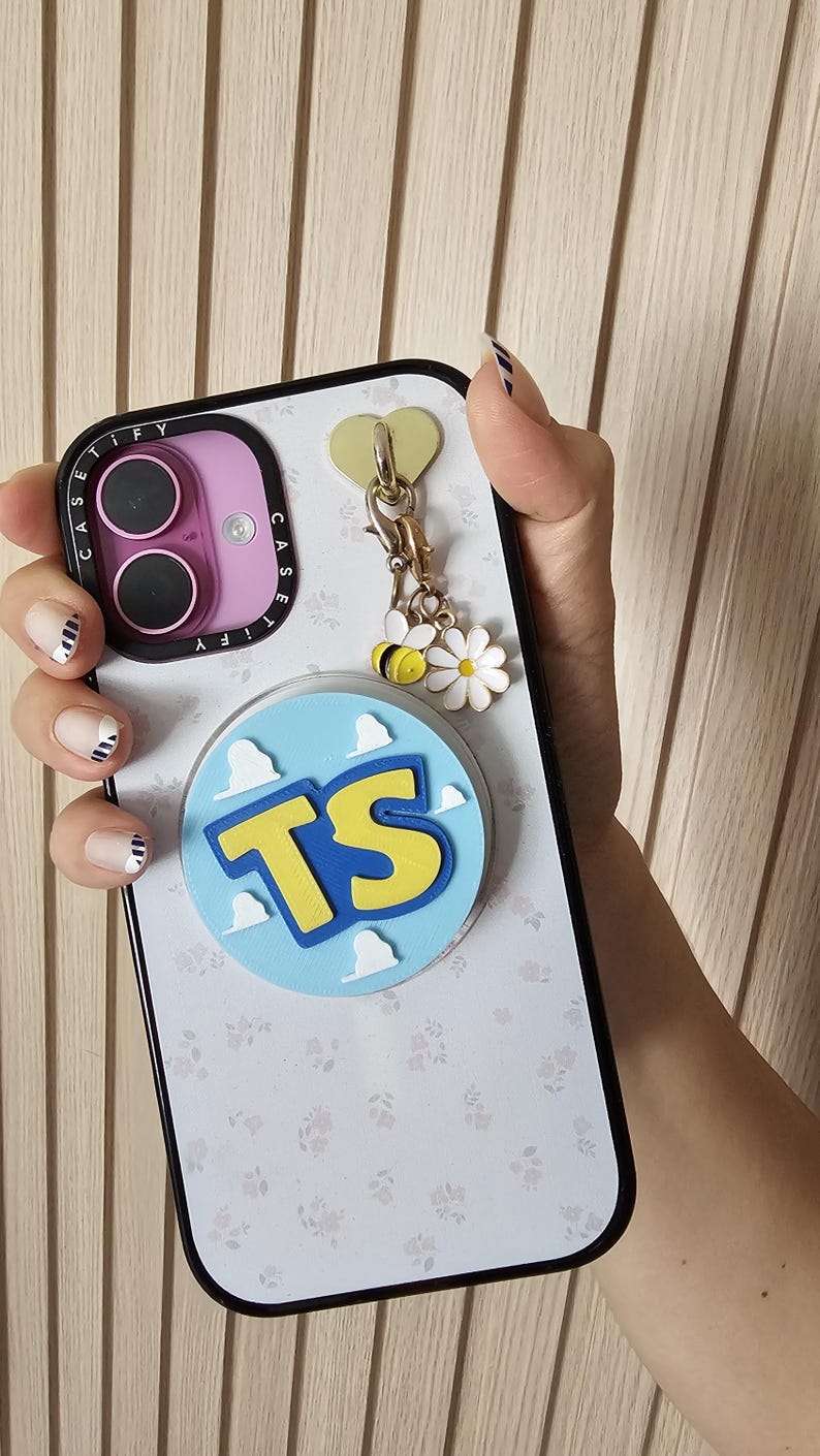 May include: A white smartphone case with a subtle floral pattern, a purple camera frame labeled CASETiFY, and a circular light blue grip featuring white clouds and the yellow letters TS. A gold heart charm hangs with a bee and a white flower.