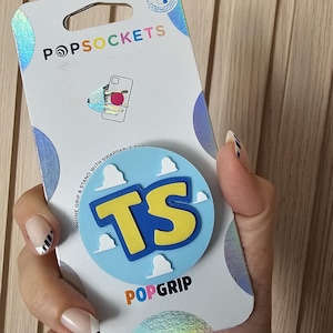 May include: A PopSockets PopGrip phone stand and grip in its original white packaging. The circular grip features a light blue background with white cloud shapes and the yellow letters TS in the center. The packaging includes the brand name PopSockets.