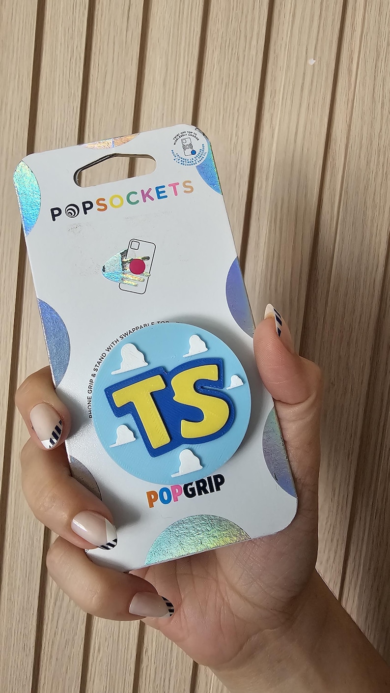 May include: A PopSockets PopGrip phone stand and grip in its original white packaging. The circular grip features a light blue background with white cloud shapes and the yellow letters TS in the center. The packaging includes the brand name PopSockets.