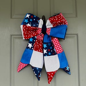 May include: A large, patriotic bow made of fabric patches in red, white, and blue. The bow features star and polka dot patterns, with a gingham check and solid blue accents. It is hanging on a gray door.
