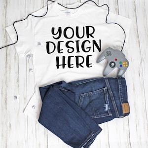 May include: A white t-shirt with the text "YOUR DESIGN HERE" printed on the front. The t-shirt is laid out on a white wooden surface with a pair of blue jeans and a grey video game controller.