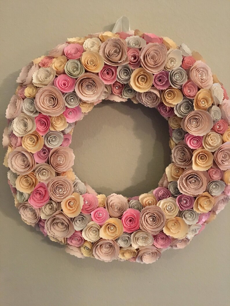 May include: A wreath made of paper flowers in shades of pink, beige, and cream. The flowers are arranged in a circular pattern and have a delicate, layered look.