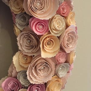 May include: A wreath made of paper flowers in shades of pink, peach, and beige. The flowers are arranged in a circular pattern and have a delicate, layered look. Each flower has a small pearl in the center.