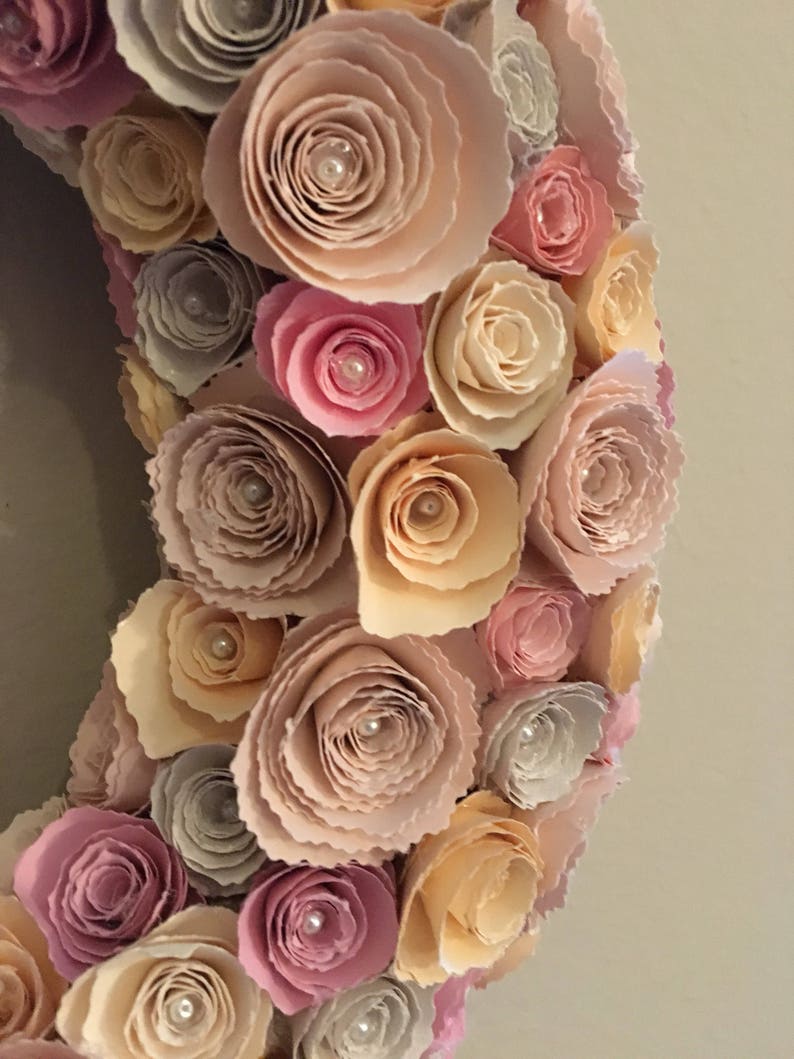 May include: A wreath made of paper flowers in shades of pink, peach, and beige. The flowers are arranged in a circular pattern and have a delicate, layered look. Each flower has a small pearl in the center.