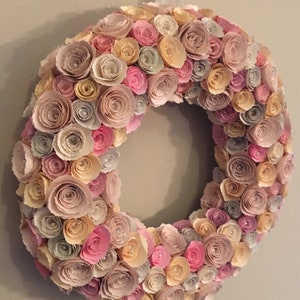 May include: A wreath made of paper flowers in shades of pink, beige, and cream. The flowers are arranged in a circular pattern and have a delicate, layered look.