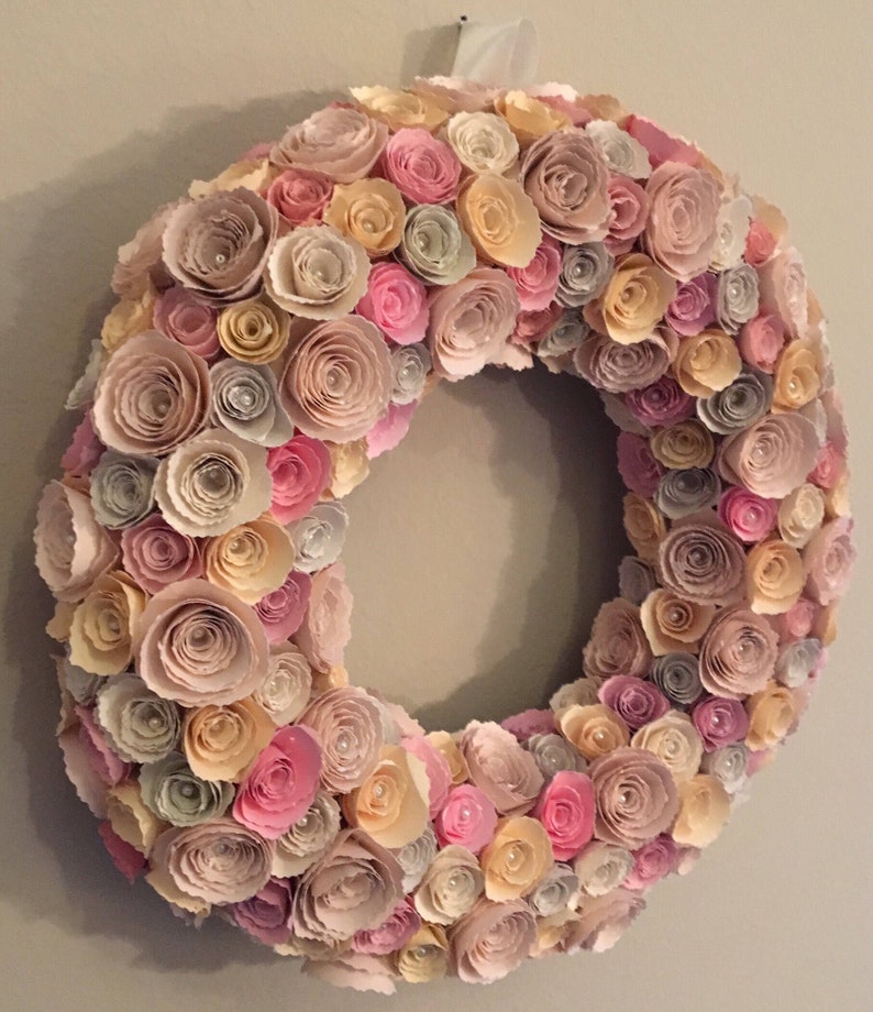 May include: A wreath made of paper flowers in shades of pink, beige, and cream. The flowers are arranged in a circular pattern and have a delicate, layered look.