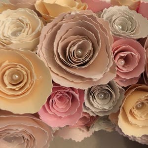 May include: A close-up of a wreath made of paper roses in shades of pink, peach, and cream. Each rose has a pearl in the center.