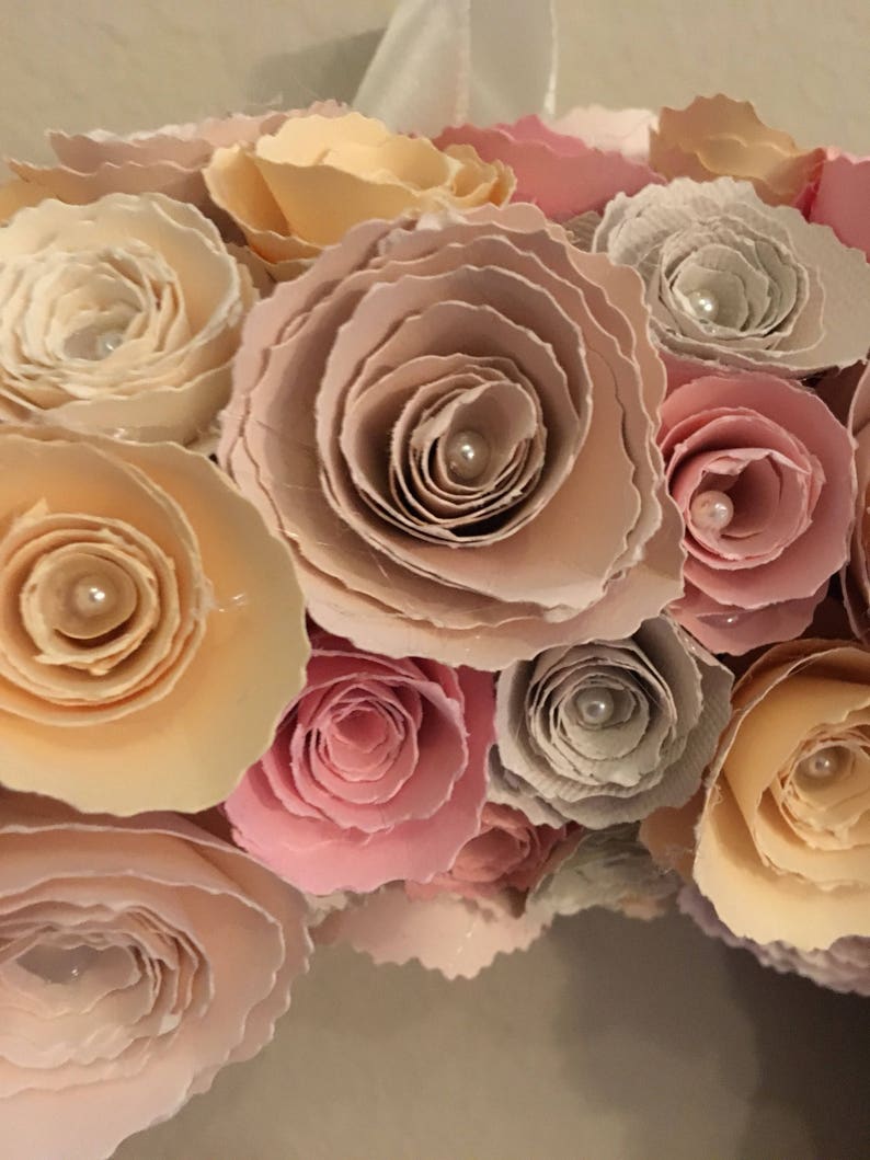May include: A close-up of a wreath made of paper roses in shades of pink, peach, and cream. Each rose has a pearl in the center.