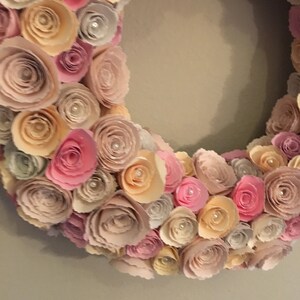 May include: A paper flower wreath with pink, peach, and cream colored flowers. The wreath is made of many layers of paper flowers, creating a full and lush look.
