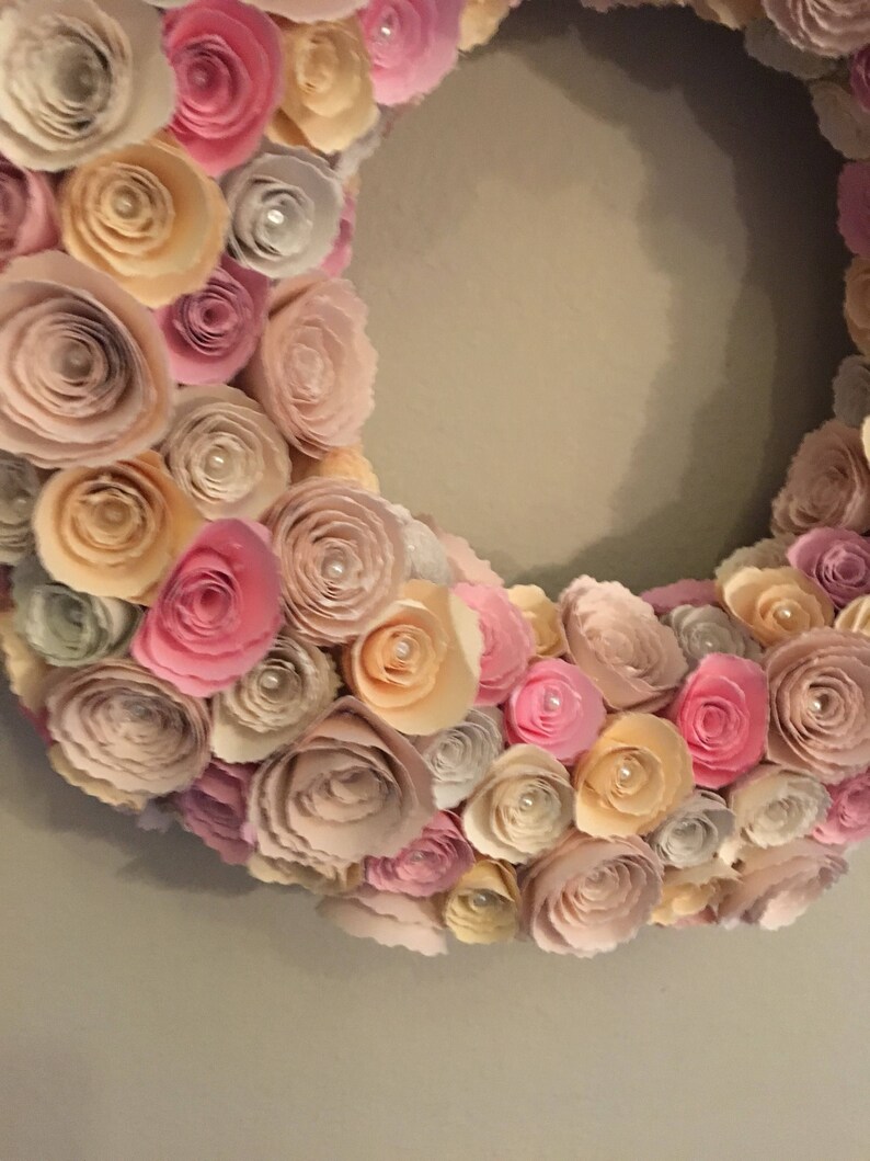 May include: A paper flower wreath with pink, peach, and cream colored flowers. The wreath is made of many layers of paper flowers, creating a full and lush look.