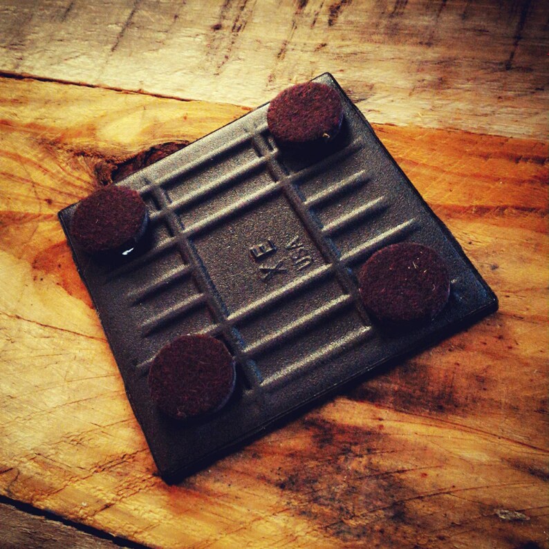 May include: Black square tile with a grid pattern and four brown felt pads on the bottom. The tile has the text "Made in USA" printed on it.