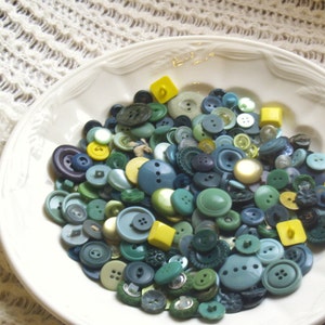 May include: A white ceramic bowl filled with a variety of blue and green buttons. The buttons are of different sizes and shapes, and some have a yellow center.