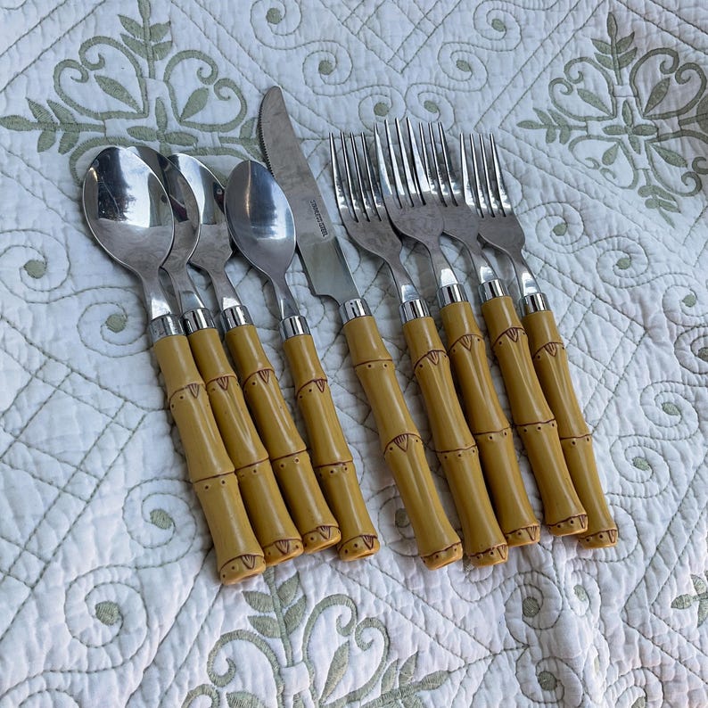 May include: A set of stainless steel flatware with bamboo-style handles. The set includes spoons, forks, and knives. The handles are a light yellow color with brown detailing. The flatware is arranged on a white patterned surface.