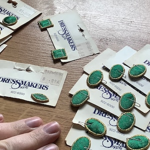 May include: Assortment of green fabric covered buttons with gold trim, packaged on cards with the text "Dressmakers Supply" and a phone number. The buttons are in various shapes, including oval, square, and leaf.