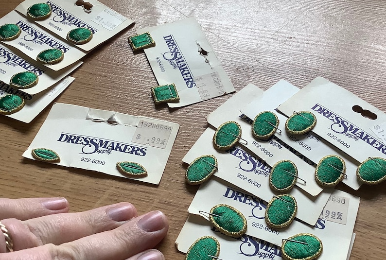 May include: Assortment of green fabric covered buttons with gold trim, packaged on cards with the text "Dressmakers Supply" and a phone number. The buttons are in various shapes, including oval, square, and leaf.