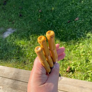 May include: Three bamboo-style, yellow-toned handles with carved details. The handles are held in a hand, with a blurred green grass background. The handles appear to be for a tool or utensil.