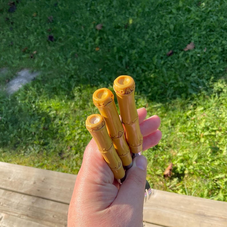 May include: Three bamboo-style, yellow-toned handles with carved details. The handles are held in a hand, with a blurred green grass background. The handles appear to be for a tool or utensil.