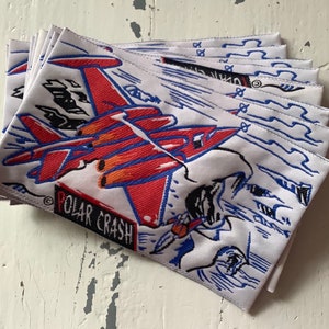 May include: A stack of white fabric patches with a red, blue, and black illustration of a jet plane with the text "POLAR CRASH" printed on the bottom.