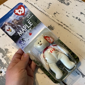 May include: White Ty Beanie Baby plush toy named "Maple the Bear" wearing a red ribbon and a Canadian flag patch. The packaging features a red and white Ty logo and the words "Collect All 4! While supplies last."