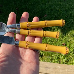 May include: A close-up of three pieces of silverware: a fork, a knife, and a spoon. Each piece has a silver head and a yellow bamboo-style handle with brown accents. The silverware is set against a green background.