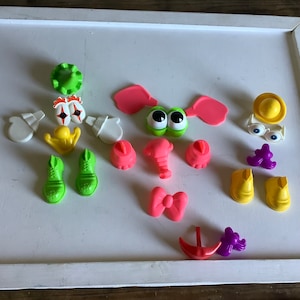 Puede incluir: Un conjunto de piezas de juguete de plástico de colores para un juego de cabeza de patata. Las piezas incluyen ojos, orejas, narices, bocas, sombreros, zapatos y una pajarita. Las piezas son de varios colores, incluyendo verde, rosa, amarillo y morado.