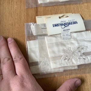 Puede incluir: Dos puños de tela de satén blanco con un delicado diseño de mariposa bordada. Los puños están empaquetados con una etiqueta que dice "Satin Cuff Dressmakers Supply 922-6000 9,75 cm 9,50 cm".