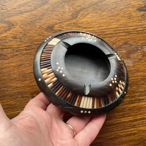 May include: A round wooden ashtray with a dark brown finish. The ashtray is decorated with a border of small, white, bone-like pieces and a ring of brown and white sticks.