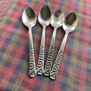 以下が含まれることがあります： 花柄とスクロール模様のディテールが施された、4本の銀色のスプーン。スプーンは、赤、緑、茶色の色合いのチェック柄の表面に配置されています。スプーンは光沢のある反射面を持っています。